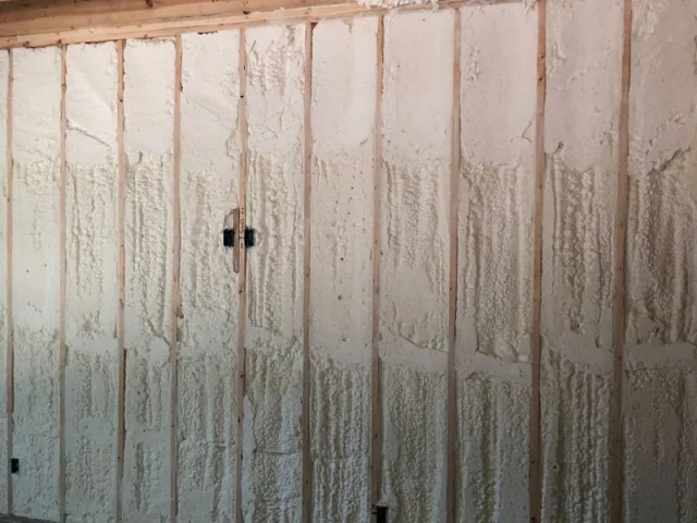Closed-cell spray foam in a framed basement wall, dense and off-white, sprayed to a partial depth that leaves the stud faces proud