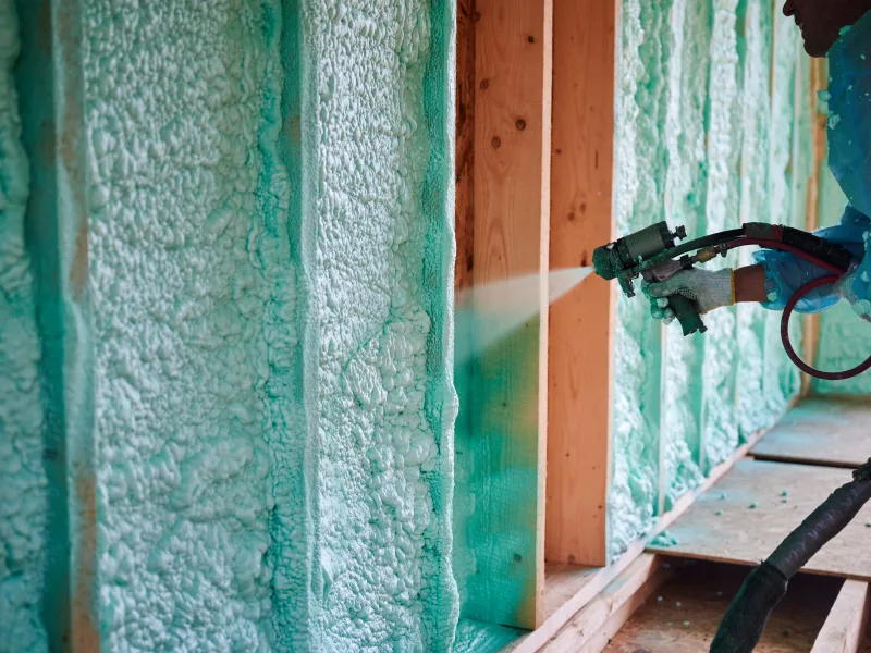 Spray gun laying foam across open stud bays along a framed wall