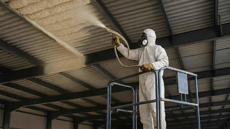 Applicator on a lift spraying a continuous layer of foam across the metal roof deck of a steel building